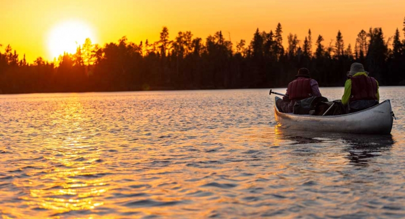 Two people sit in a canoe on calm water as the sun sets behind trees on the shore. 